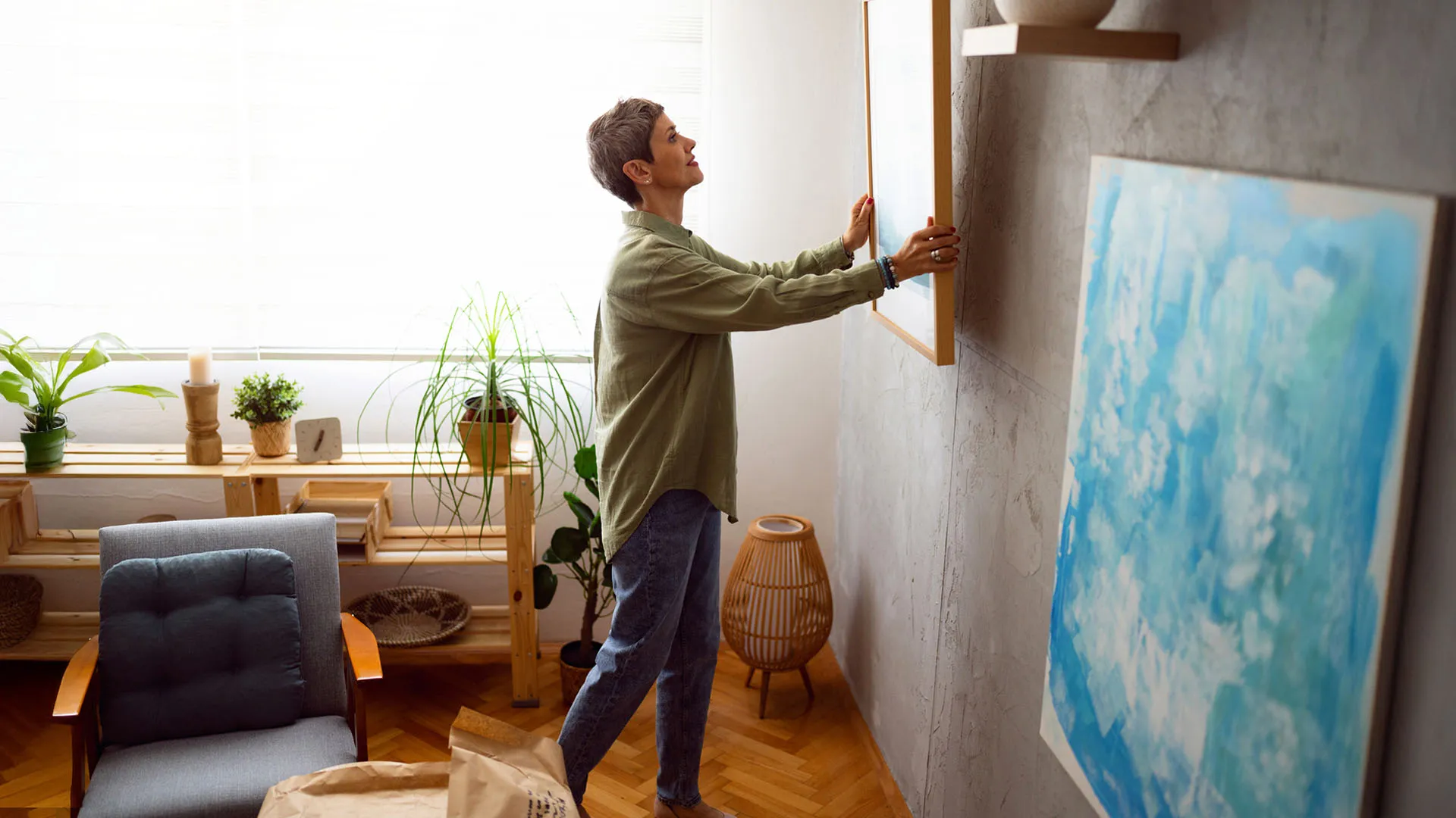 Person hanging a framed picture on a gray wall in a cozy room with plants and a gray cushioned chair.